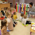 Powiększ zdjęcie Dzieci siedzą na podłodze i słuchają pani czytającej bajkę