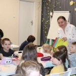 Powiększ zdjęcie Pani Zosia stoi przy stoliku przy którym siedzą dzieci w ręku trzyma kolorowe kartki