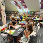 Powiększ zdjęcie Dzieci na warsztatach w bibliotece siedzą przy stolikach i wykonują pracę plastyczną