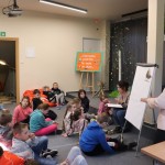 Powiększ zdjęcie Dzieci siedzą na podłodze w oddziale dla dzieci słuchają opowiadania bibliotekarki