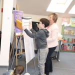 Powiększ zdjęcie Chłopiec wraz z bibliotekarką przykleja pasek zadania na flipczart