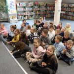 Powiększ zdjęcie Dzieci siedzą na podłodze w bibliotece pani czyta książkę